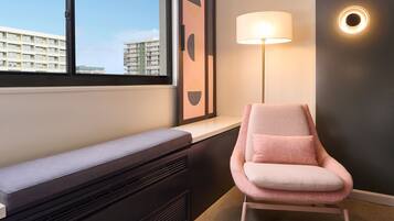 A modern room with a bench, a pink chair, a floor lamp, and a view of buildings through a window.