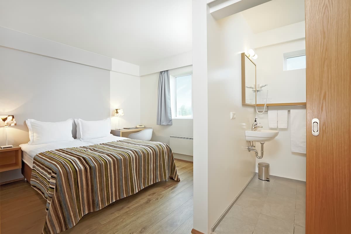 A hotel room with a bed, bedside table, lamp, and a view of the bathroom.