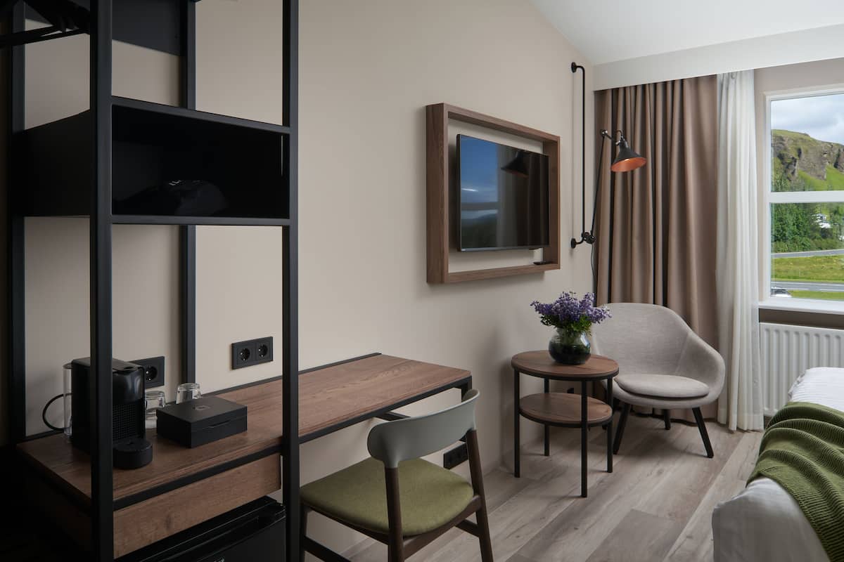 A hotel room with a wooden desk, a chair, a small table with a vase, a bed with a green blanket, a television on the wall, and a window with curtains.