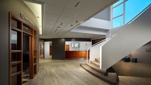 Un hall d’hôtel moderne avec un comptoir d’accueil en bois, un escalier et une grande fenêtre laissant entrer la lumière naturelle.
