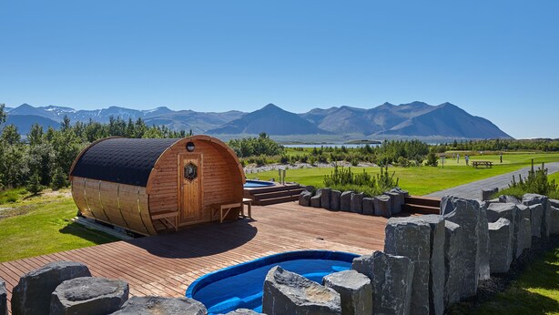 A wooden cabin with a pool and a deck, surrounded by a stone wall, set against a mountainous backdrop.