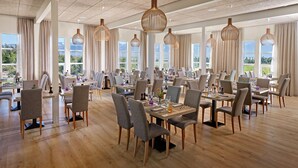 A spacious dining area with multiple tables and chairs, large windows with curtains, and hanging pendant lights.