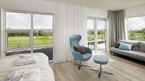 A hotel room with a bed, a blue armchair, a grey sofa, and large windows offering a view of a golf course.