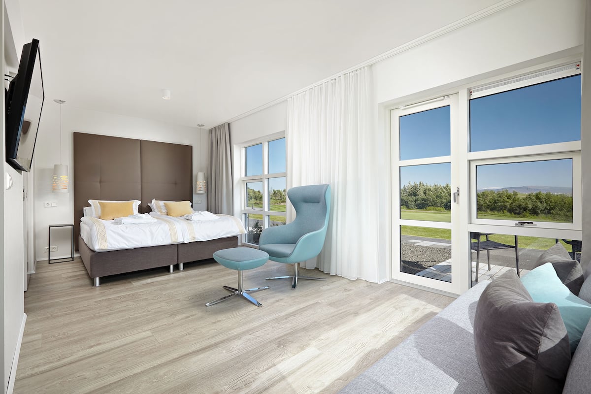 A modern hotel room with a large bed, a seating area with a chair and ottoman, and a sofa. There is a television mounted on the wall and a large window offering a view of the outdoors.