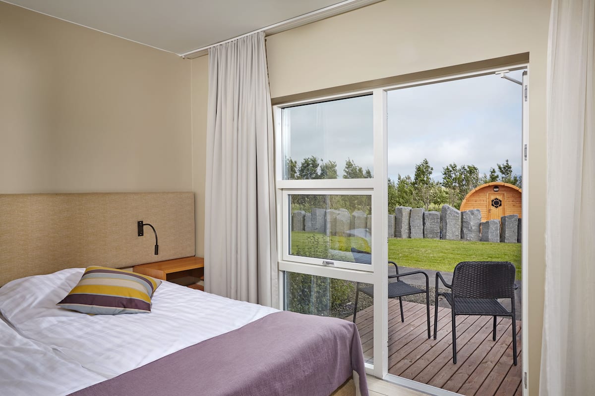 A bedroom with a bed, a nightstand, a window, and a view of an outdoor deck with chairs and a wooden structure.