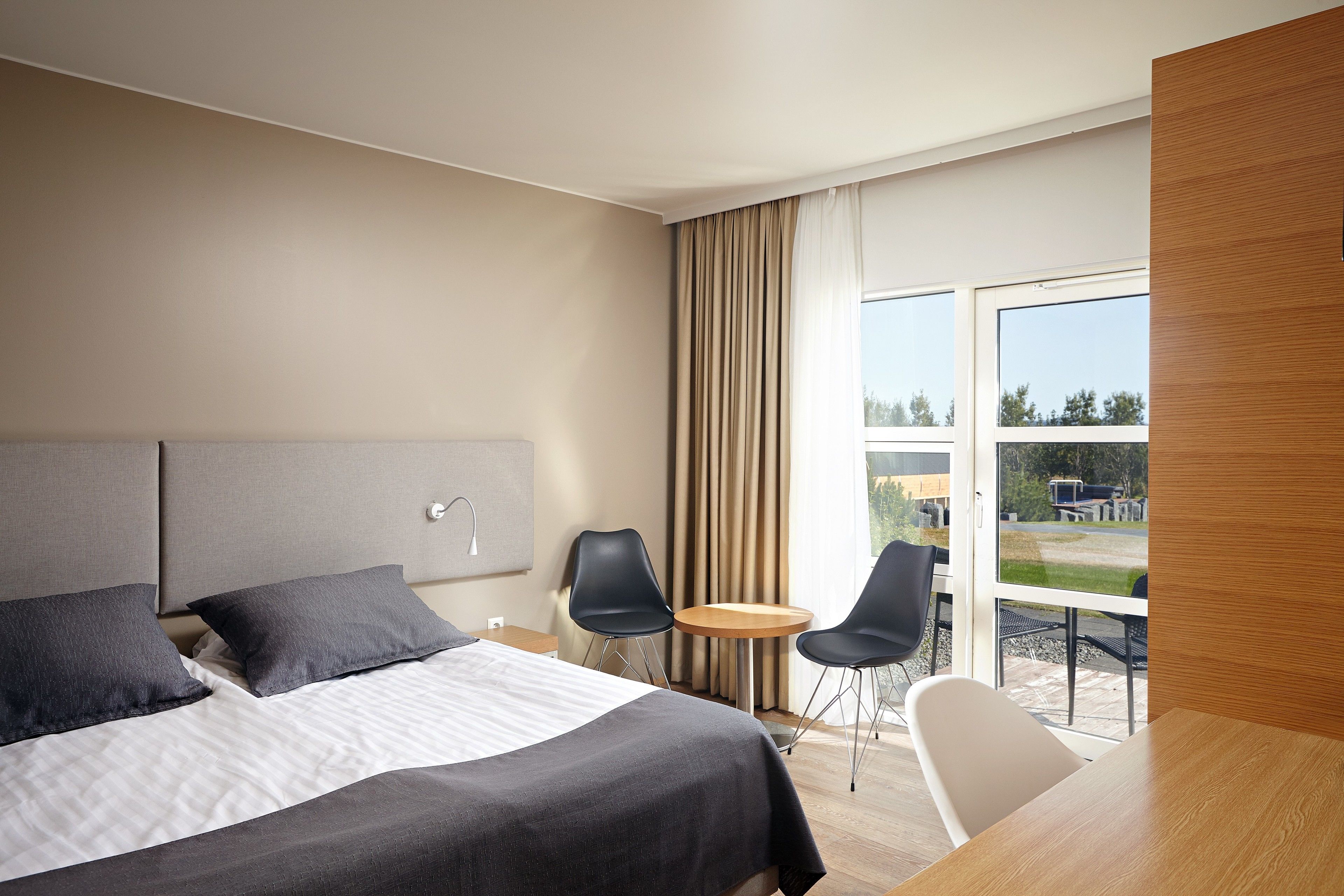 Quarto de hotel moderno com uma cama grande, duas cadeiras, uma mesa pequena e vista para o exterior.