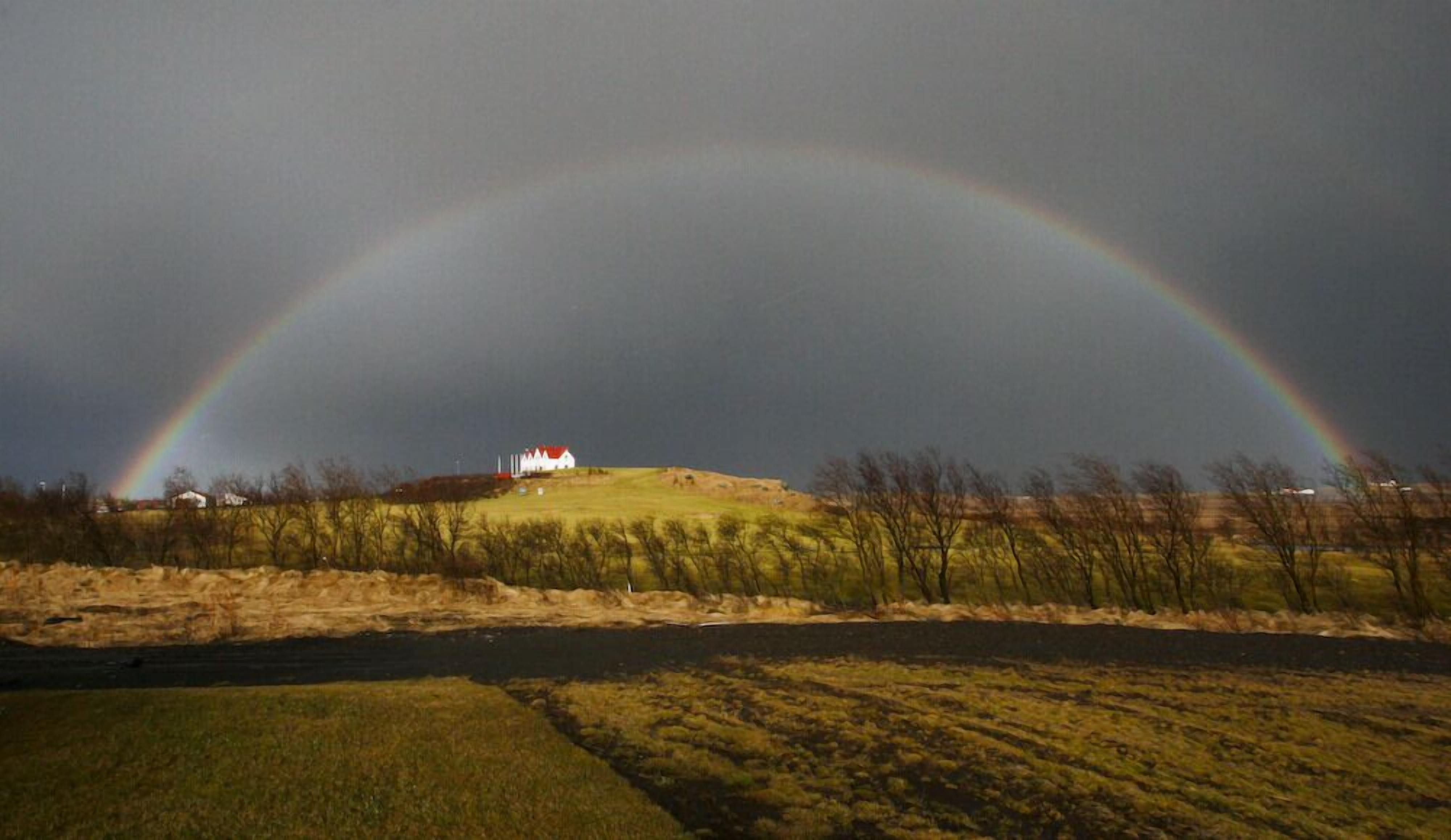 Ländliche Landschaft mit einem weißen Haus auf einem Hügel, einem Regenbogen und einem Feld.