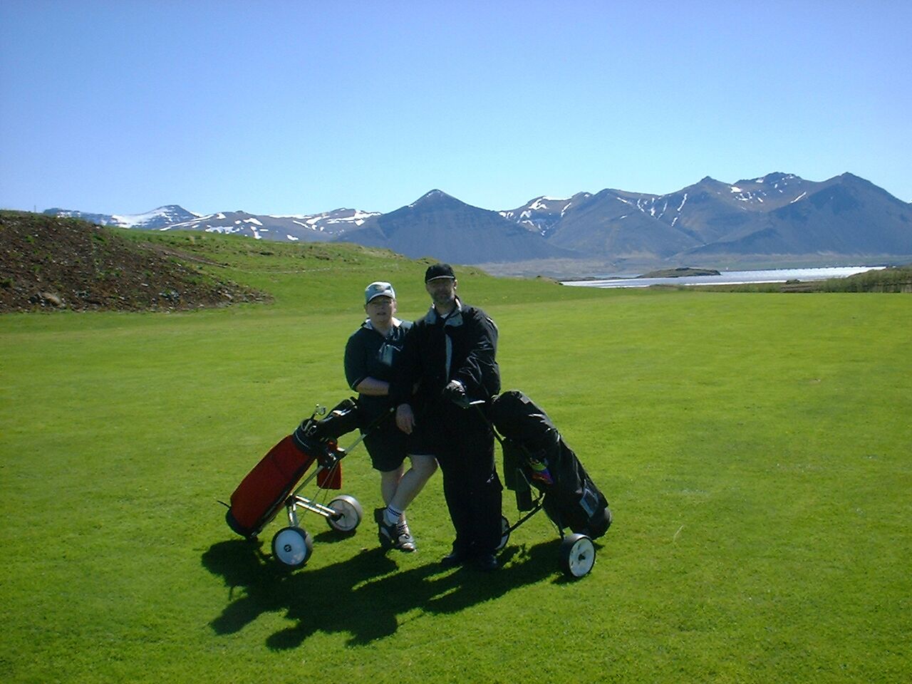 Dos personas con una tabla de snowboard y un cochecito en un campo césped con montañas al fondo.