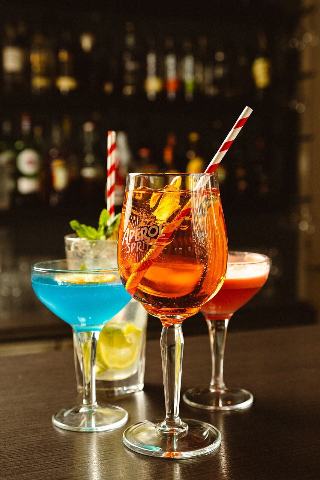 Three cocktails in martini glasses with straws, one with a red drink and another with a blue drink, on a bar counter with a blurred background of bottles.