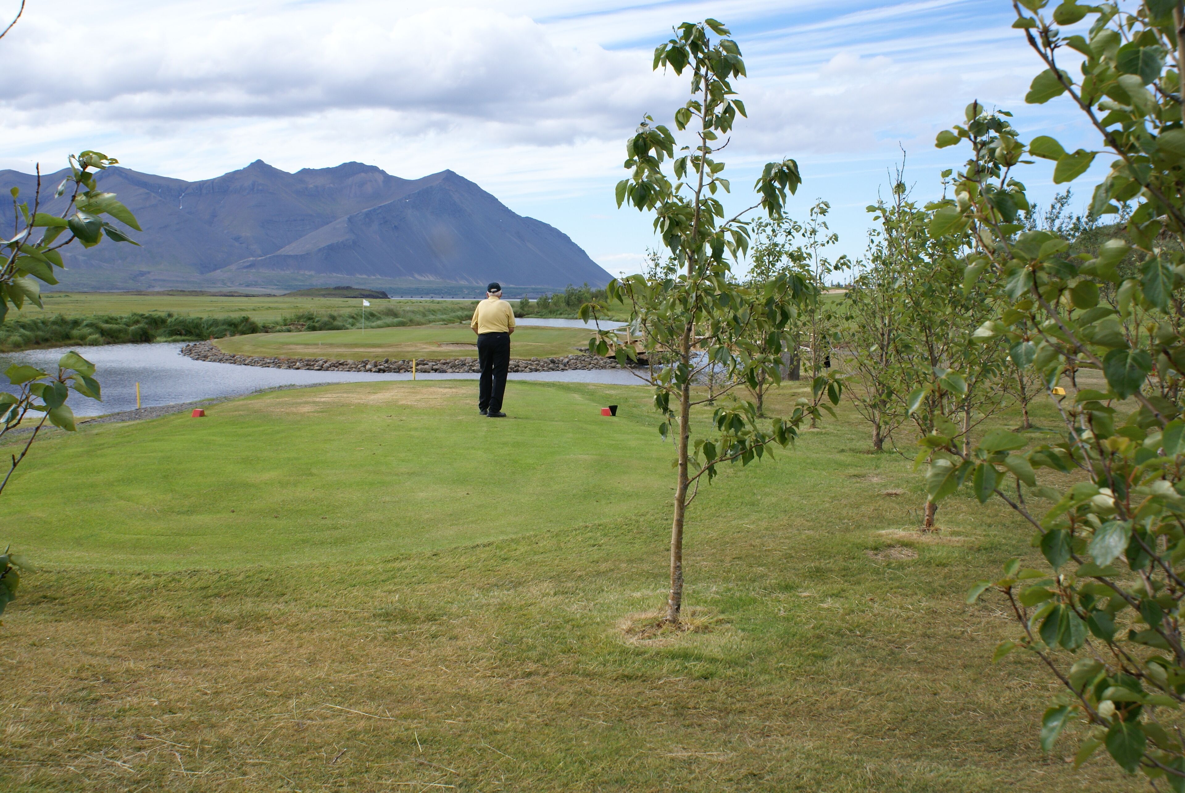 A golf course with a person standing on the green, a river, and mountains in the background.