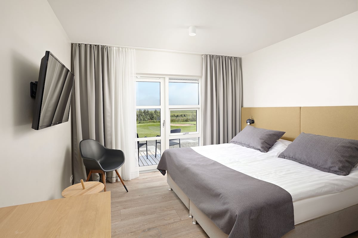 A modern hotel room with a large bed, a flat-screen TV mounted on the wall, a small table, a chair, and a balcony with a view of the outdoors.