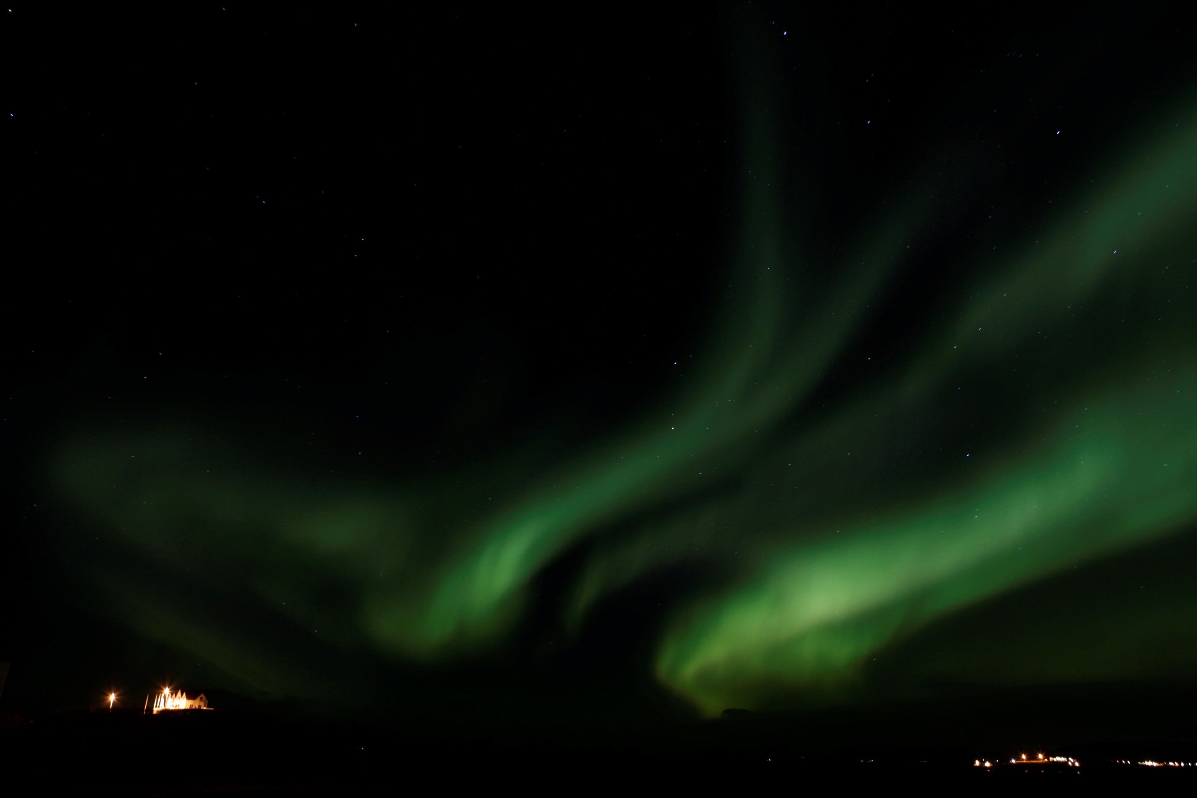 The Northern Lights over a dark landscape with distant lights in the background.