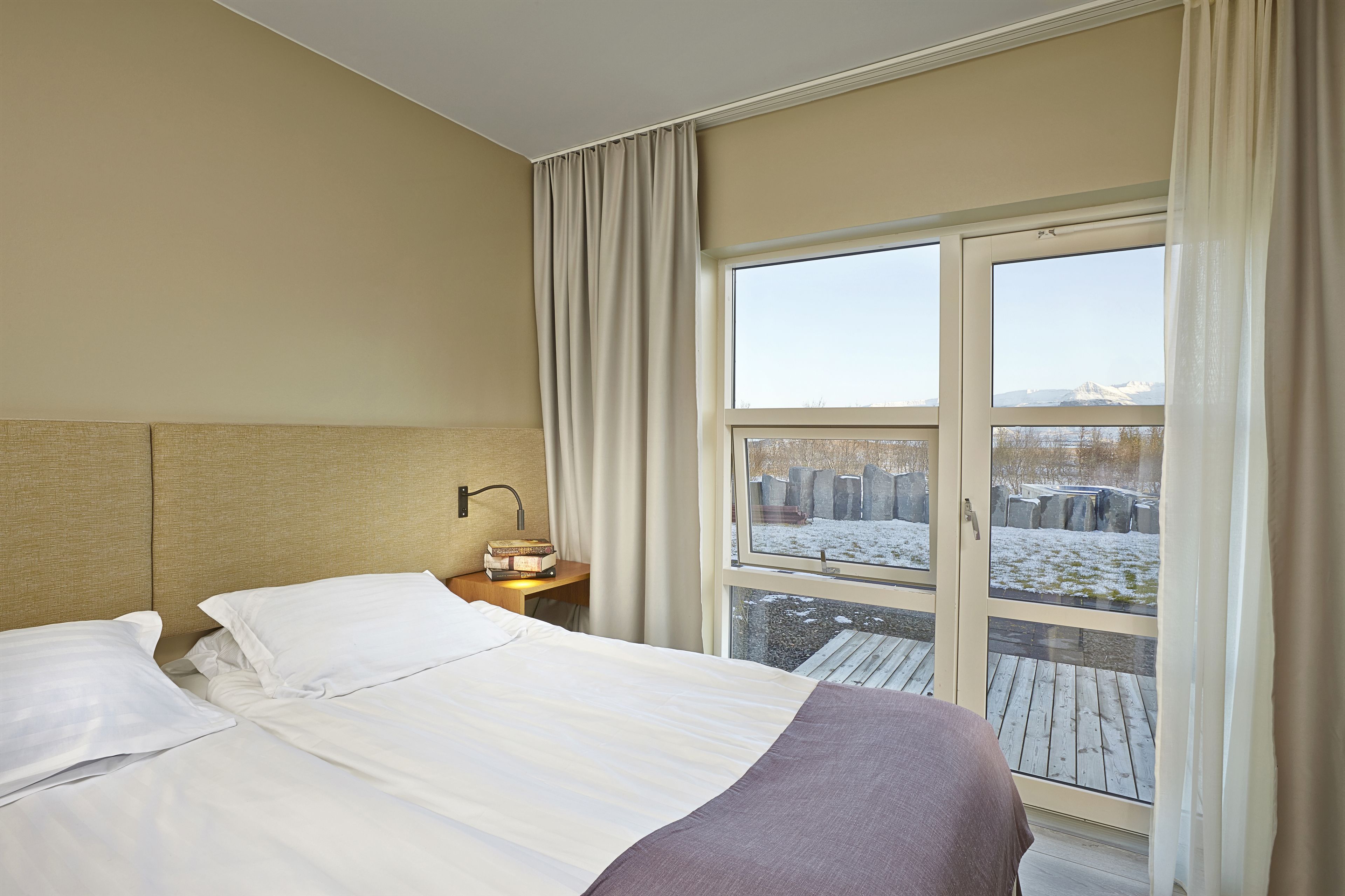 Une chambre d’hôtel avec un lit, une table de chevet, une lampe et une fenêtre donnant sur des pierres recouvertes de neige.