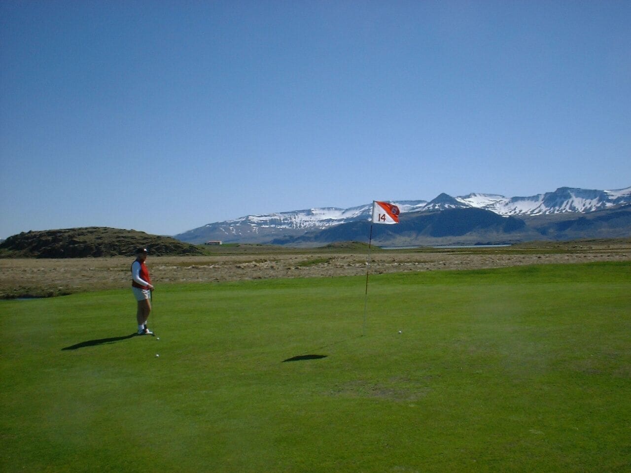 A person on a golf course with a flag marked '14' and snow-capped mountains in the background.