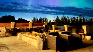 An outdoor seating area with chairs and tables, a stone wall, and a view of the night sky with the aurora borealis.