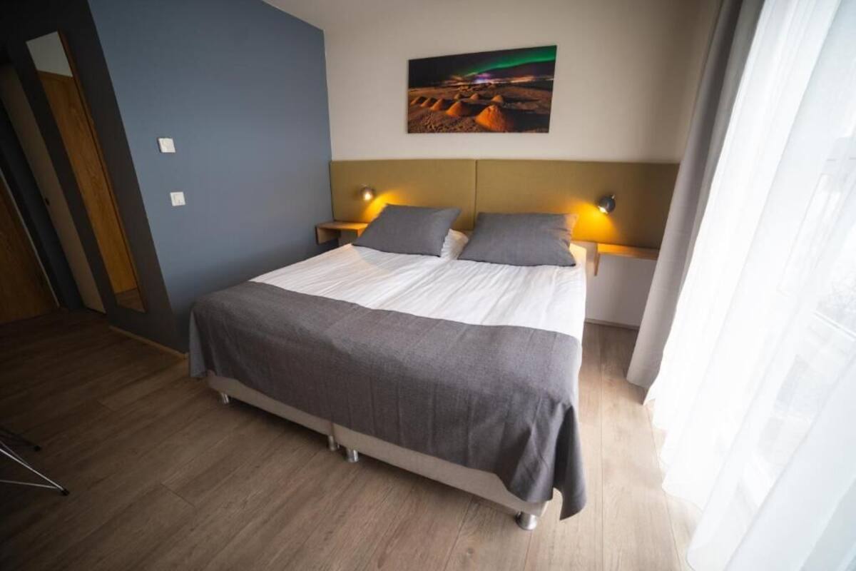 Habitación de hotel con cama, dos mesitas de noche con lámparas, una mesita auxiliar, un cuadro en la pared y una ventana con cortinas.
