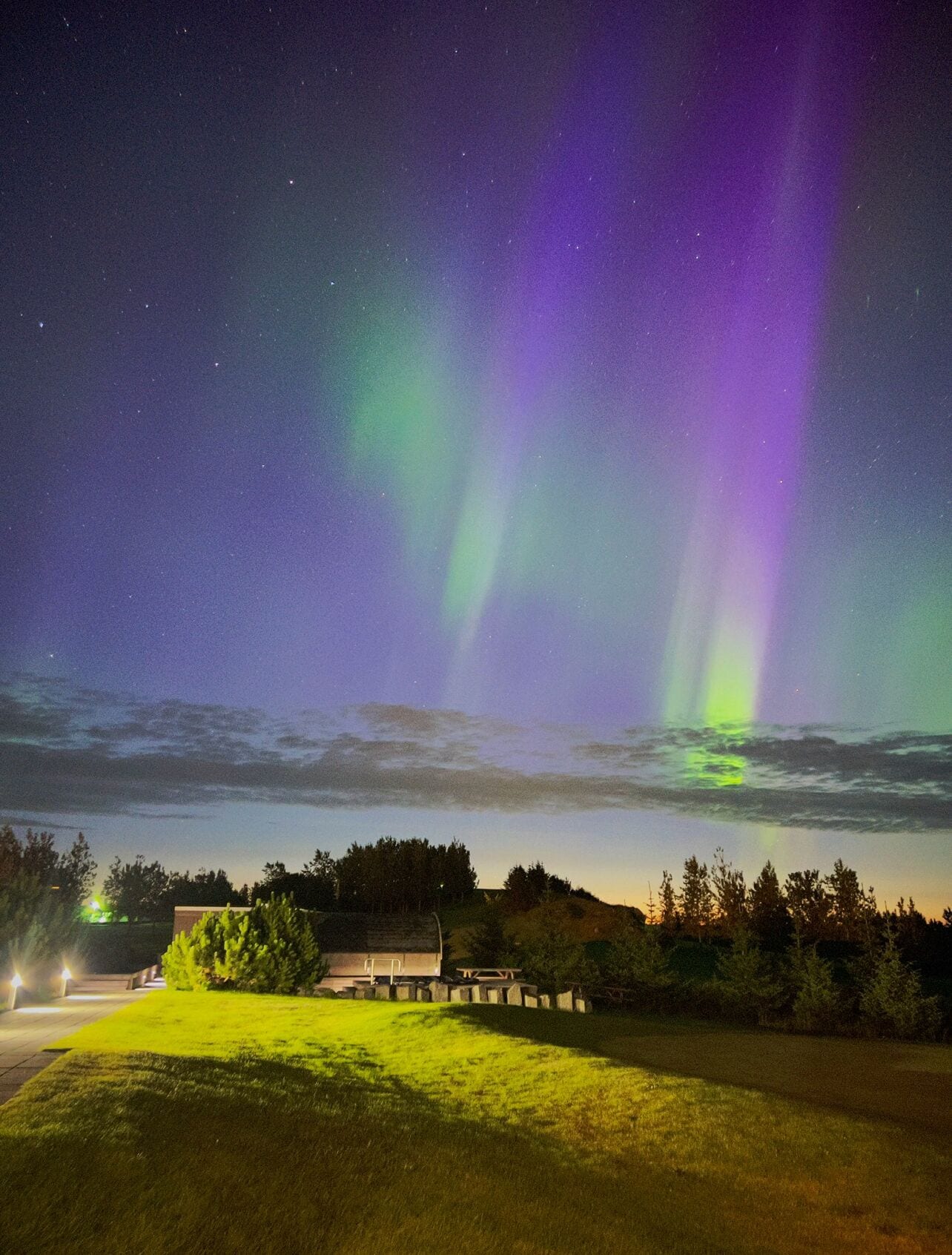 A nighttime view of the Northern Lights over a grassy field with a building and trees in the foreground.