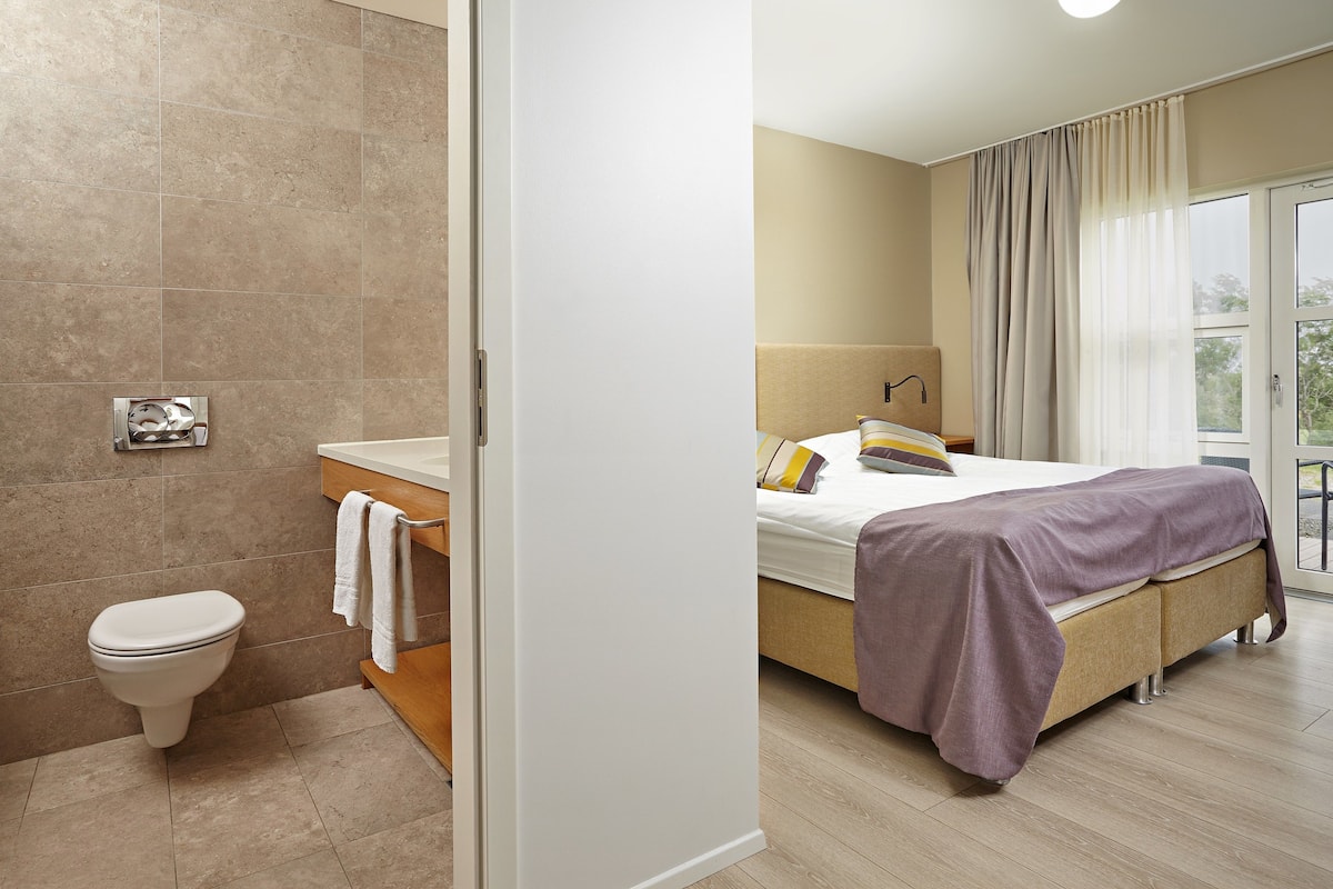 A modern hotel room with a bathroom, a bed with a purple blanket, and a view of the outdoors.