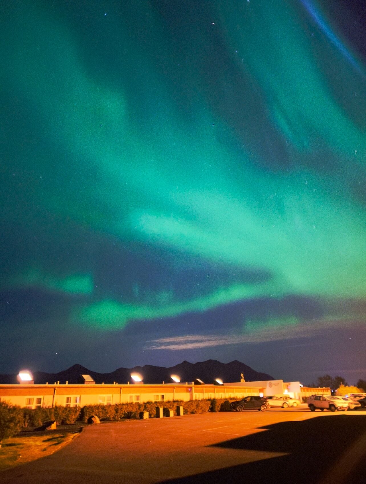 A parking lot with a building in the background and a night sky with green aurora lights.