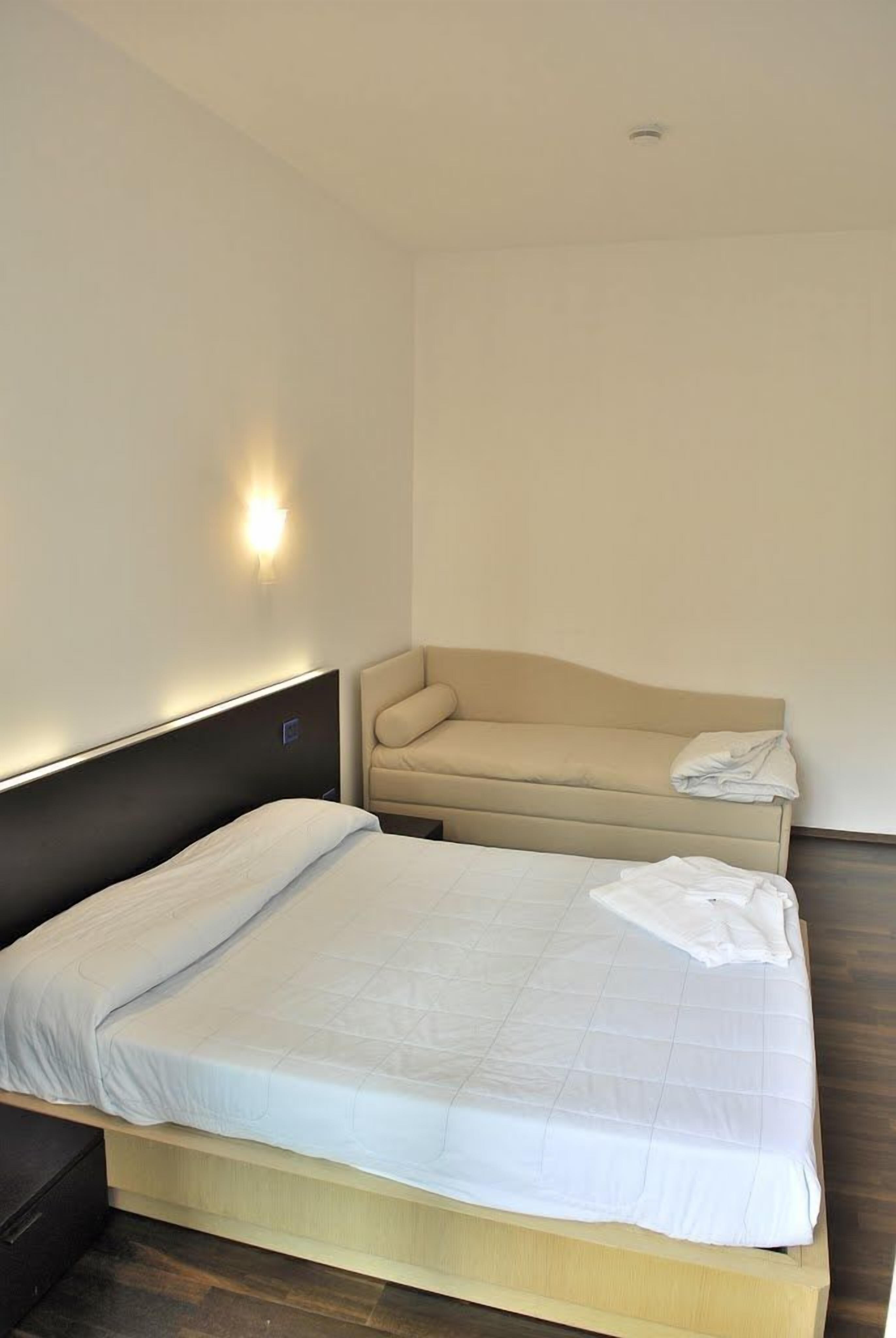 A hotel room with a bed, a bedside table, a chair, and a wall-mounted light.