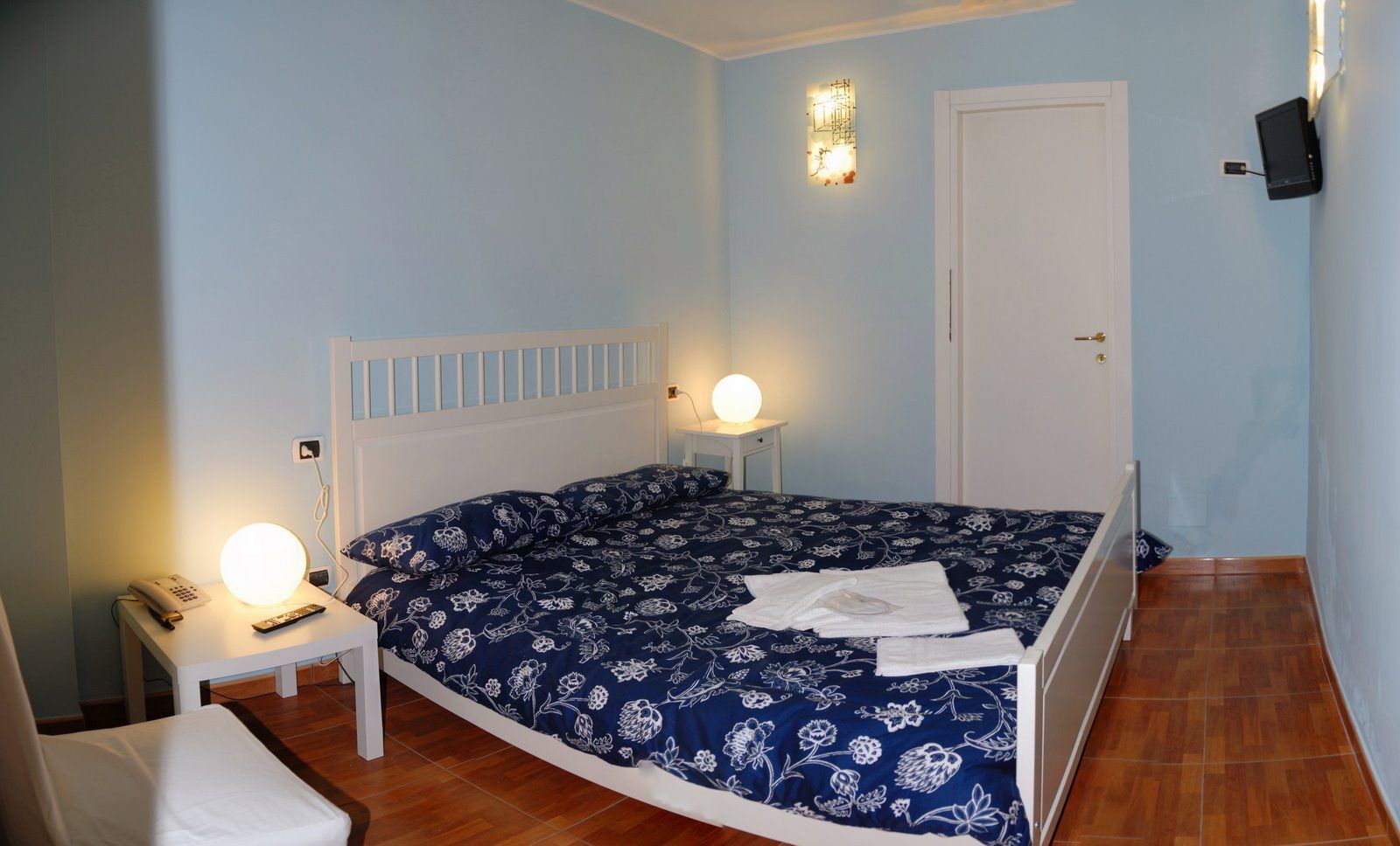 Un dormitorio con cama, mesitas de noche, un televisor montado en la pared y una puerta.