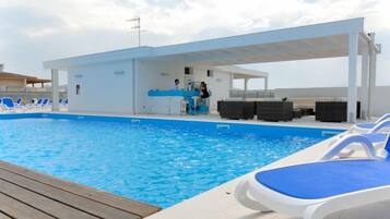 Zona de piscina con sillones de descanso, un bar y un espacio de estar cubierto.