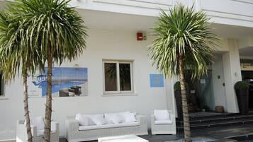 A hotel exterior with a white facade, a white wicker sofa set, two palm trees, and a poster of a pool.