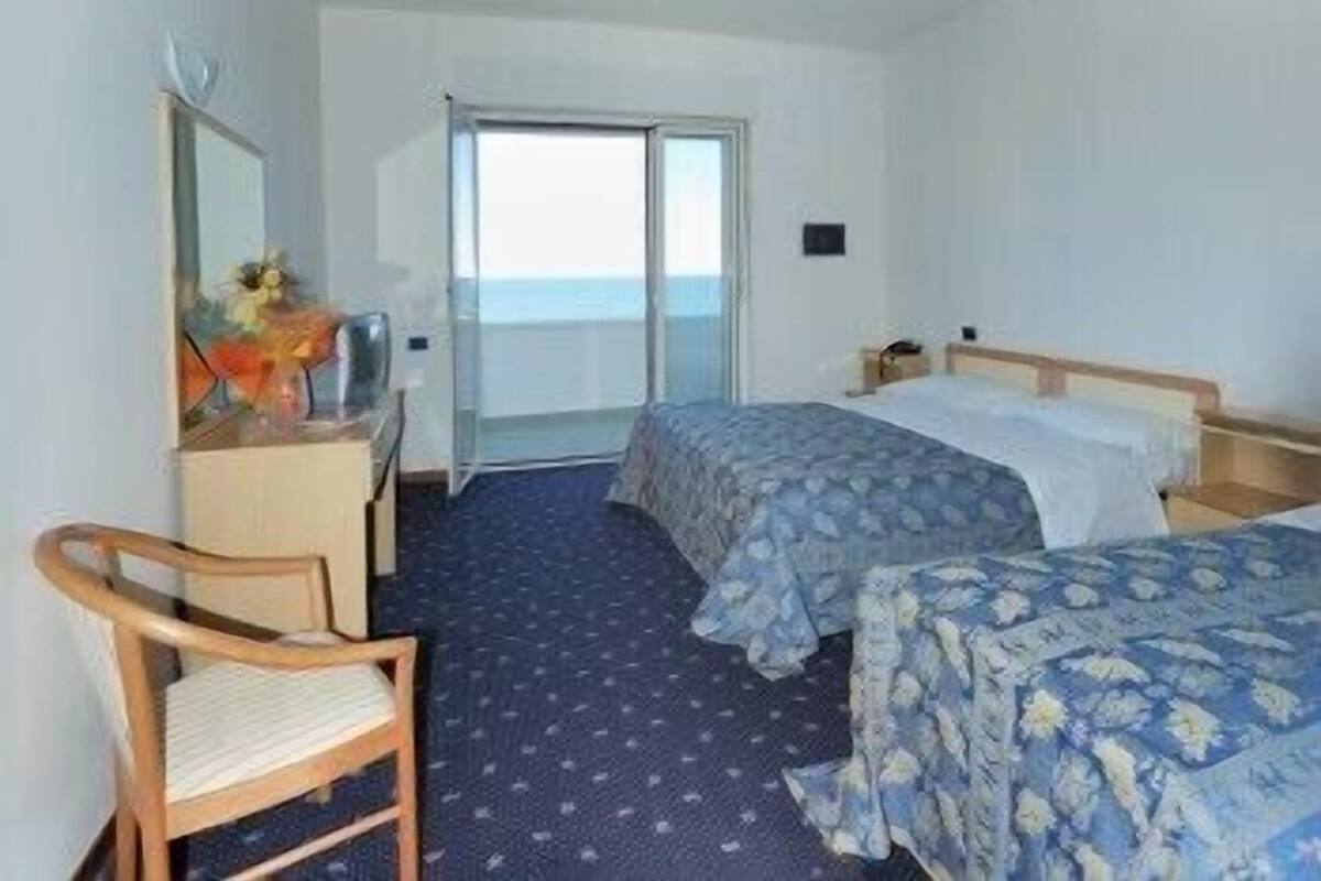 A hotel room with two beds, a desk, a chair, and a large window offering a view of the sea.
