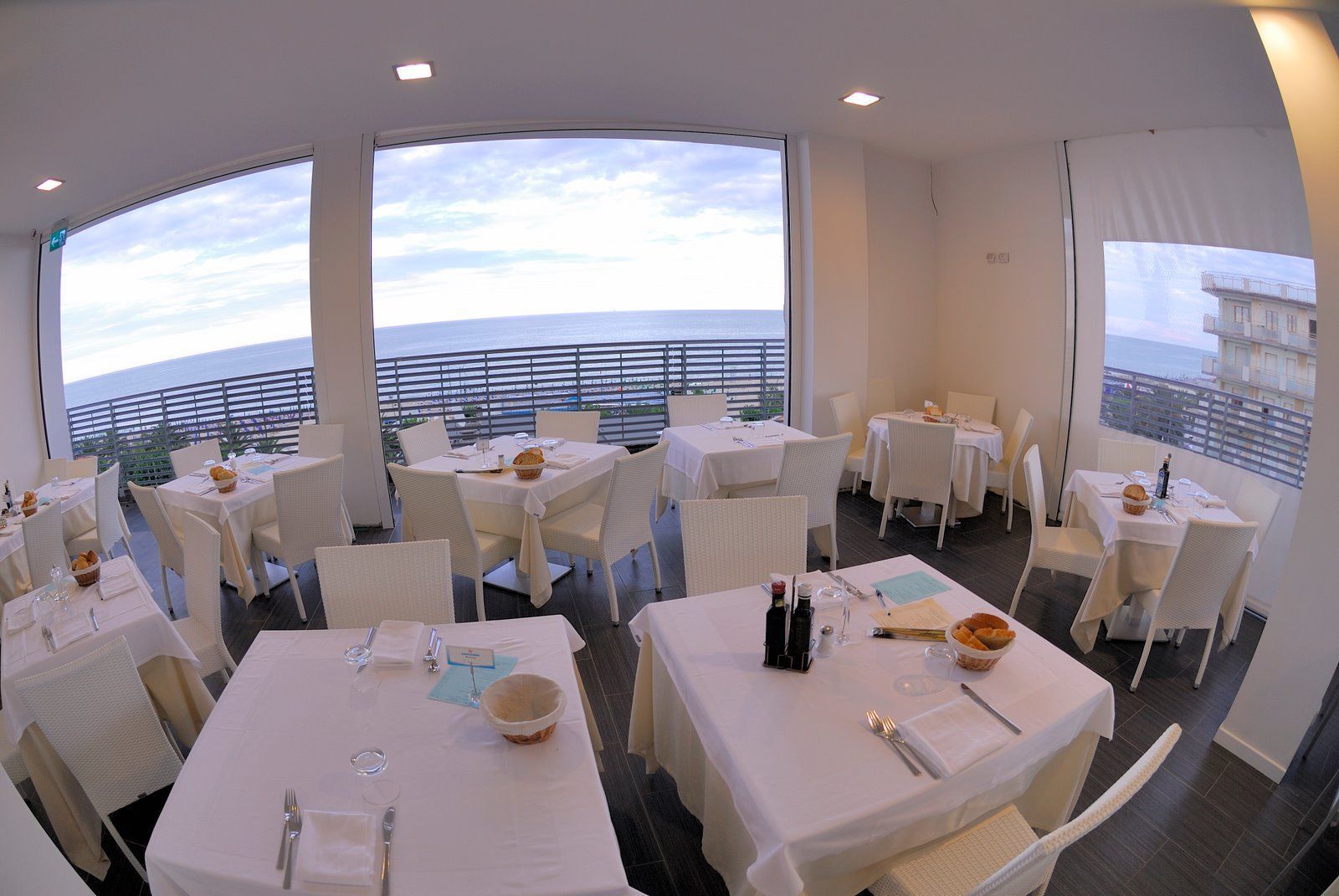 A restaurant with multiple tables set for dining, large windows offering ocean views, and a balcony with a railing.