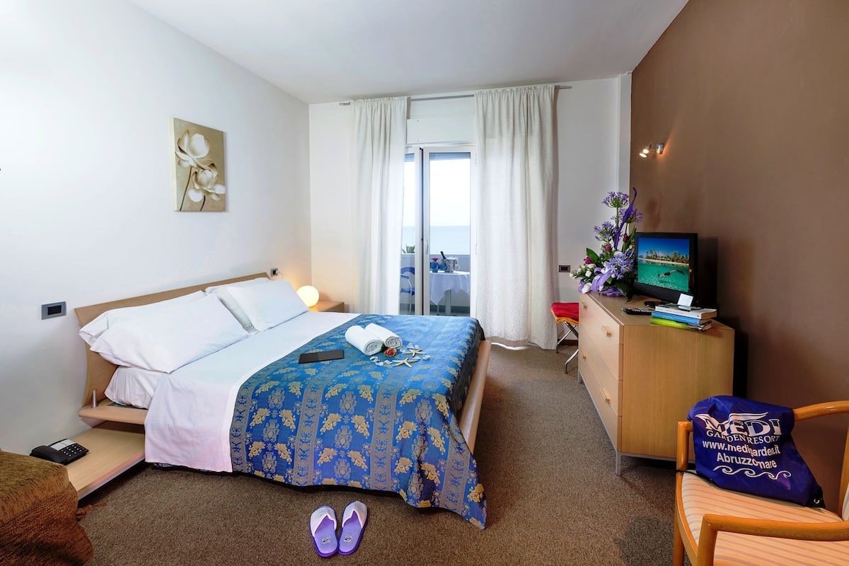 A hotel room with a bed, a TV, a desk with a computer, and a view of the sea through the window.