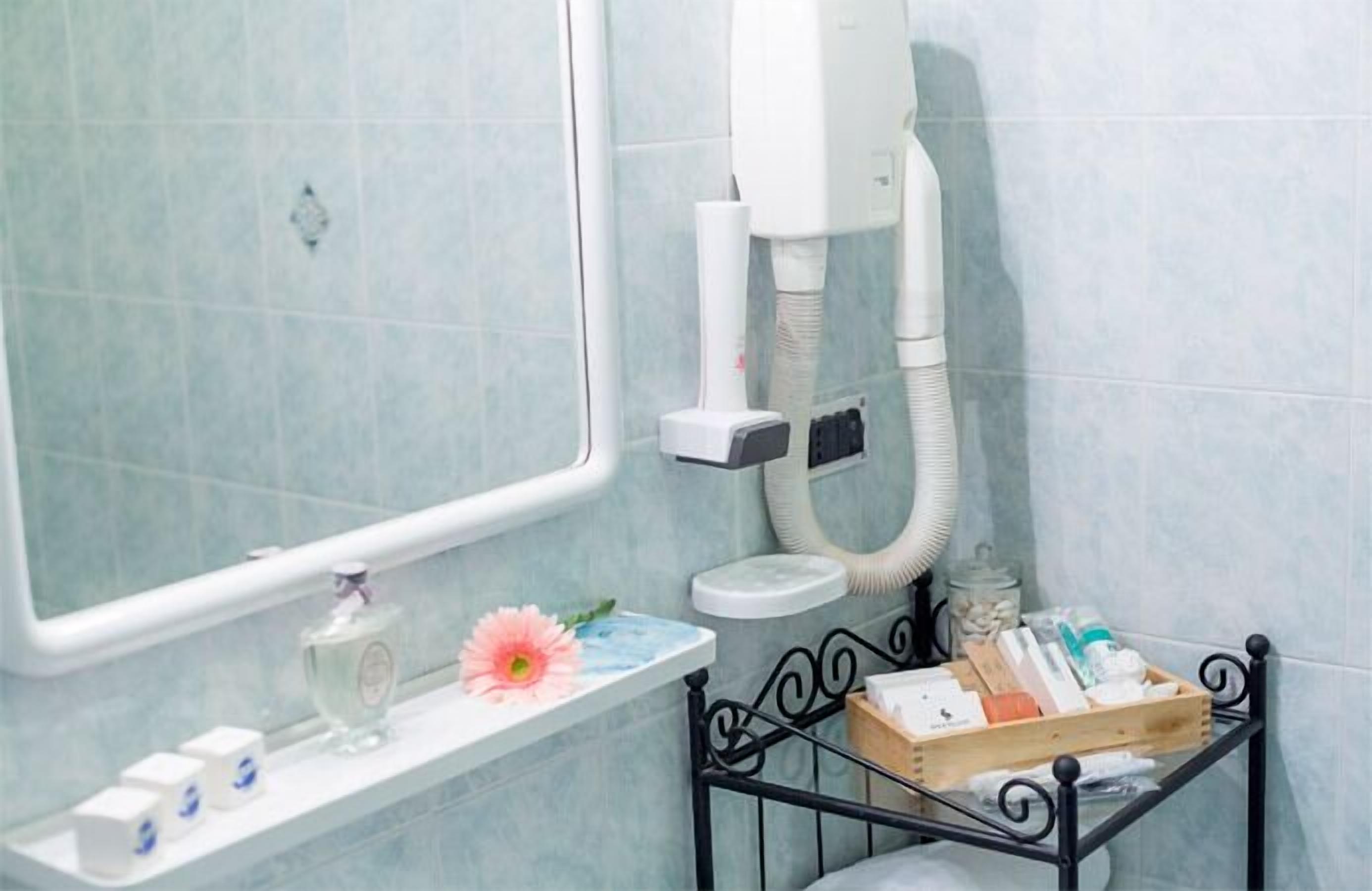 A bathroom with a mirror, a sink, a hairdryer, and toiletries on a shelf.