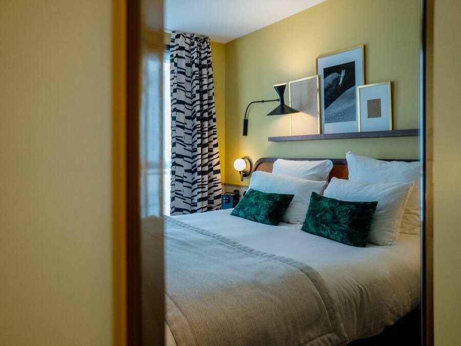 Et hotelværelse med en seng, et natbord, en lampe og en vægmonteret hylde med billeder i rammer.
