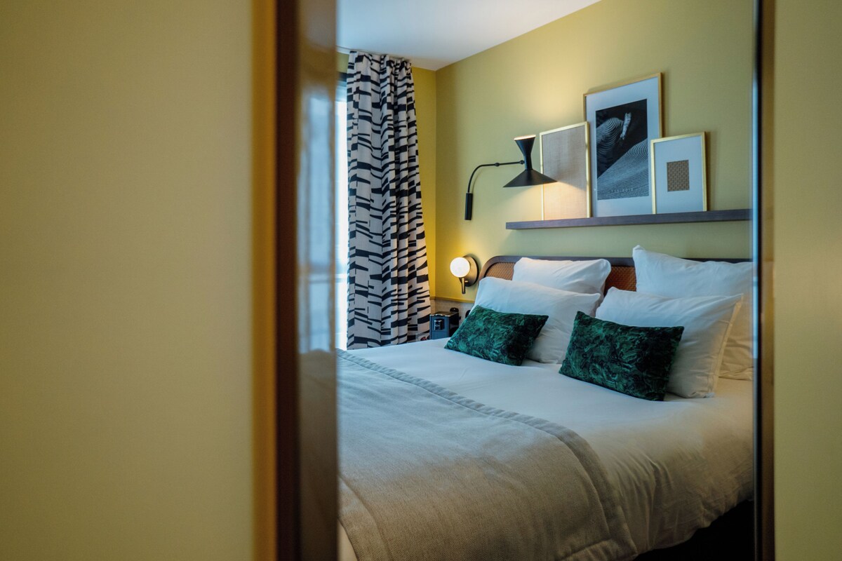 Un lit bien fait, avec des oreillers blancs et verts, un rideau à motifs noir et blanc et une lampe fixée au mur.