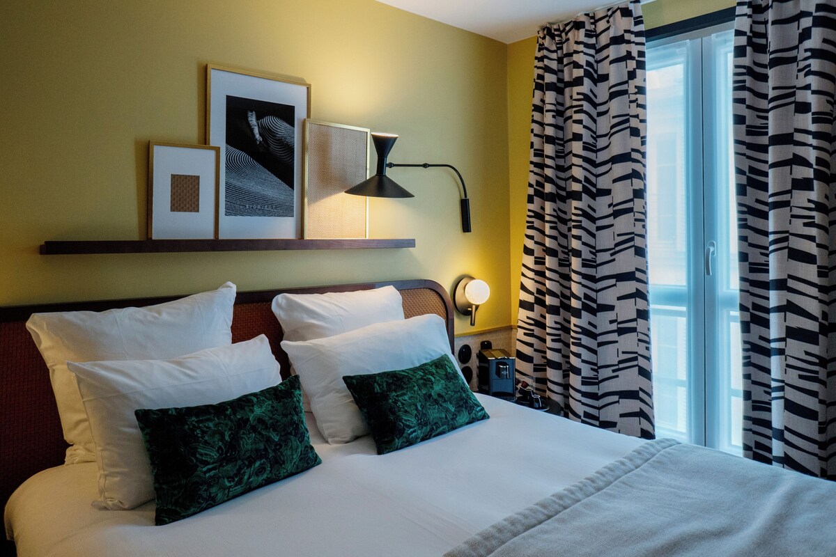Una habitación de hotel con una cama, almohadas, una mesita de noche con lámpara, cuadros en la pared y una ventana con cortinas estampadas.
