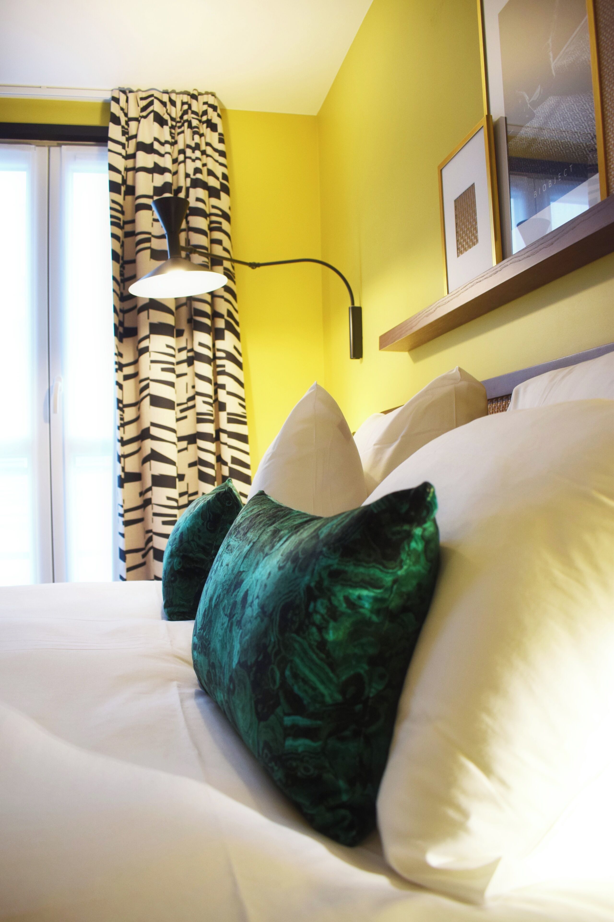 A hotel room with a bed, pillows, a lamp, a window with curtains, and a framed picture on the wall.
