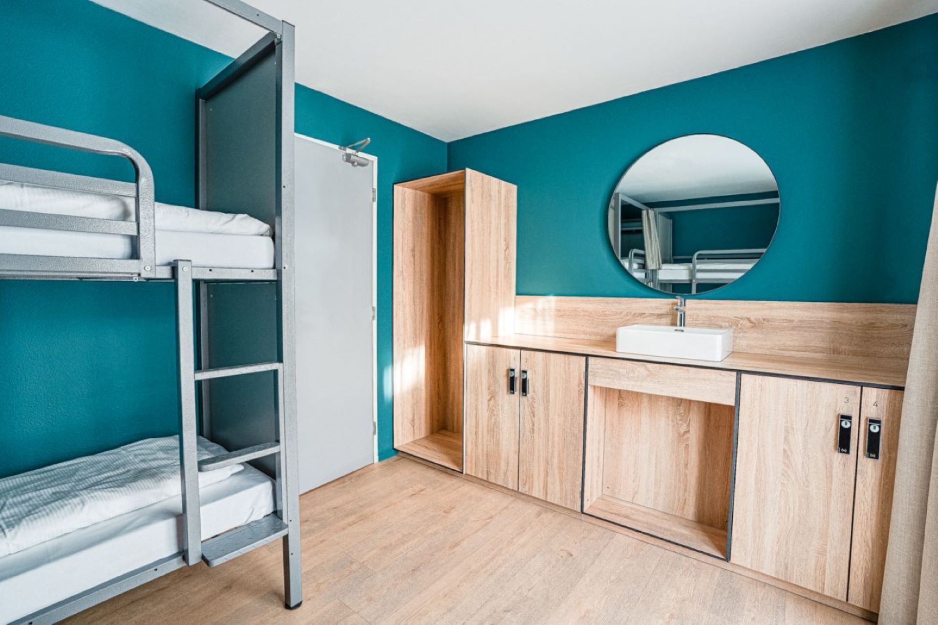 Una cama individual con estructura metálica y ropa de cama blanca, un mueble de madera con lavamanos y un espejo redondo sobre el lavamanos.