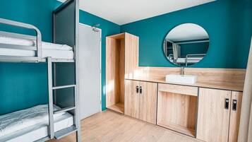 Una cama individual con estructura metálica y ropa de cama blanca, un mueble de madera con lavamanos y un espejo redondo sobre el lavamanos.