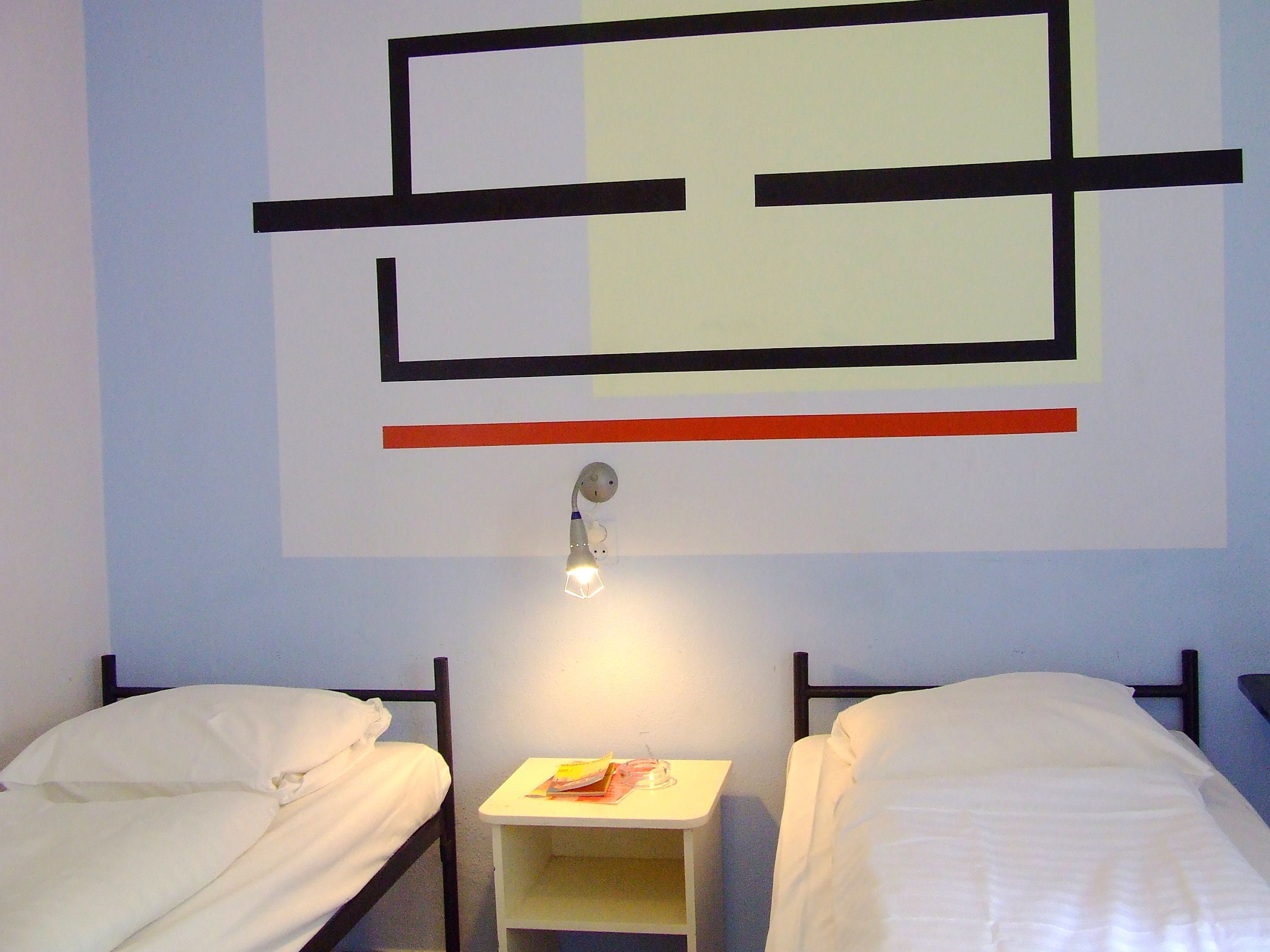A room with two single beds, a small bedside table with a lamp, and a wall with abstract geometric art.