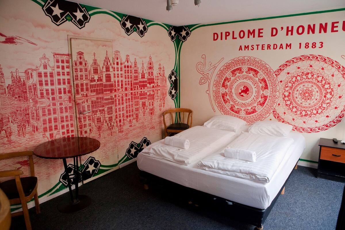 A hotel room with a bed, a wooden chair, a small round table, and a wall with red and white patterns and text.