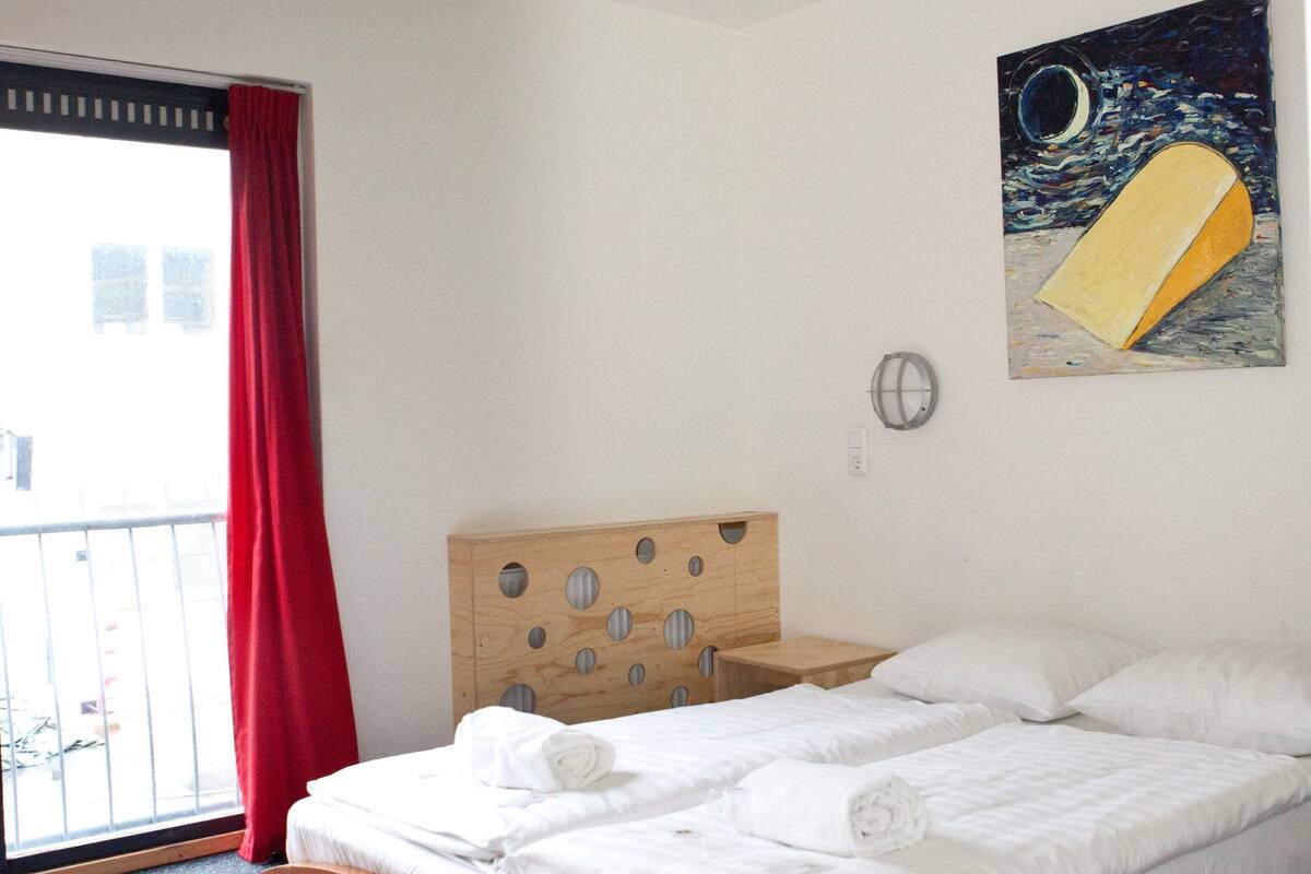 A hotel room with a wooden bed, a red and white pendant light, a painting of a boat on the wall, and a view of the outside through a window with red curtains.