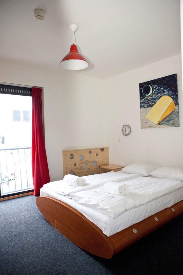 A hotel room with a wooden bed, a red and white pendant light, a painting of a boat on the wall, and a view of the outside through a window with red curtains.