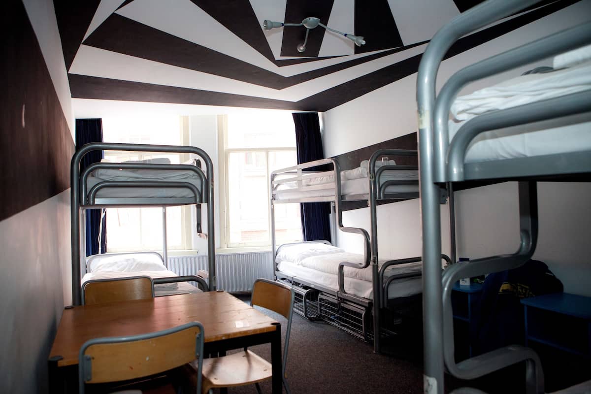 A dormitory room with bunk beds, a table, chairs, and a window providing natural light.