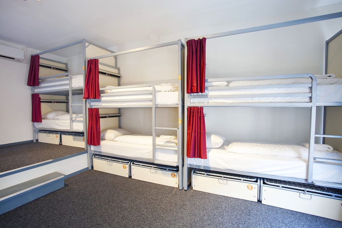 A dormitory room with bunk beds, each with a red curtain, and a small storage drawer under the bed.