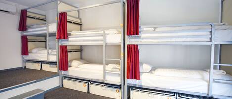 Habitación tipo dormitorio con literas, cada una con una cortina roja, y un pequeño cajón para guardar objetos debajo de la cama.