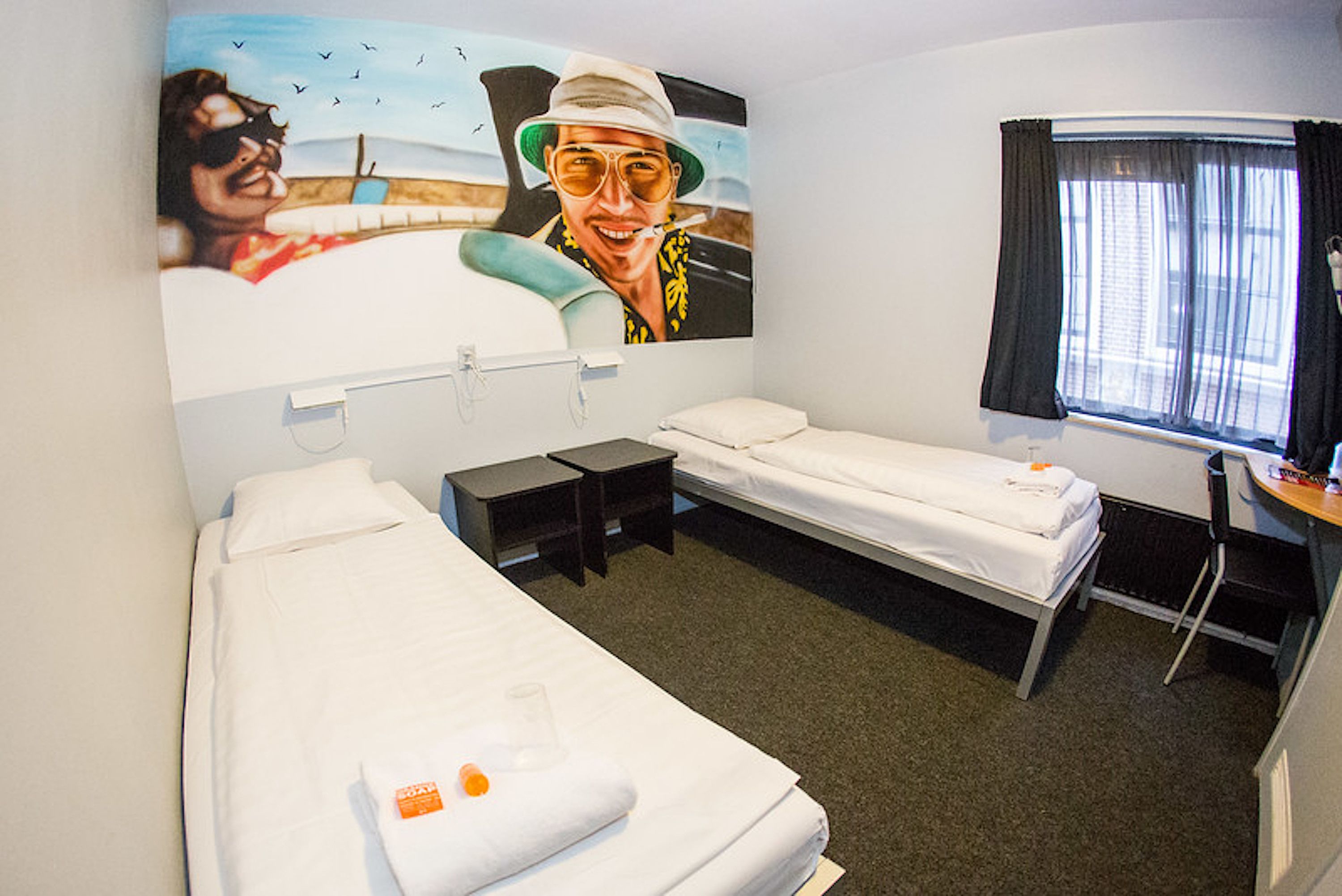 A hotel room with two single beds, a small desk, a chair, a window with curtains, and a wall mural.