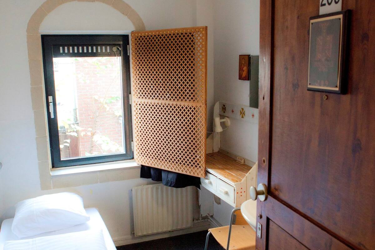 A small, compact room with a bed, a desk, and a chair. There is a window with a view of the outside, a radiator under the desk, and a door with the number 209.