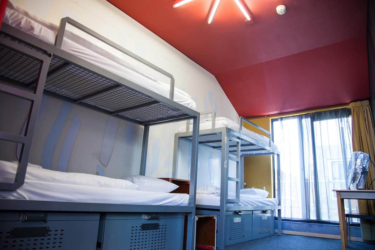 A dormitory room with bunk beds, a window with curtains, and a table with a fan.