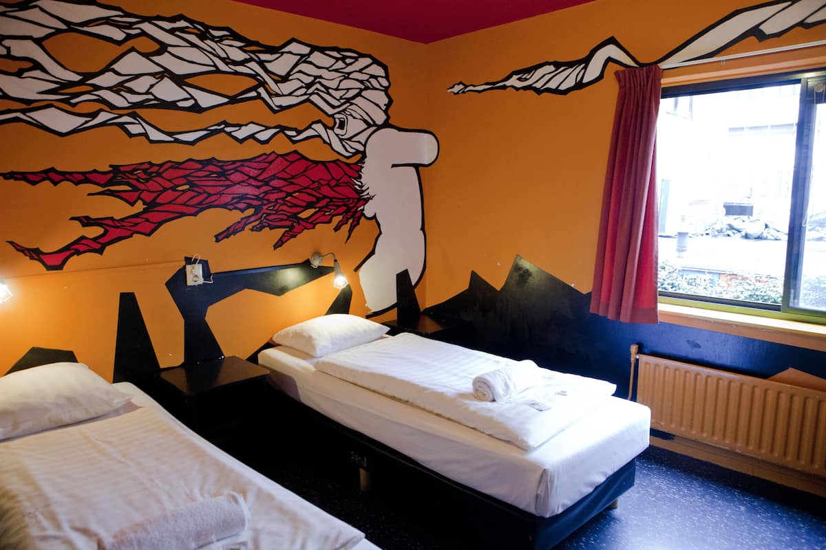 A hotel room with two beds, a mural on the wall, a window with a view of the outside, and a radiator.