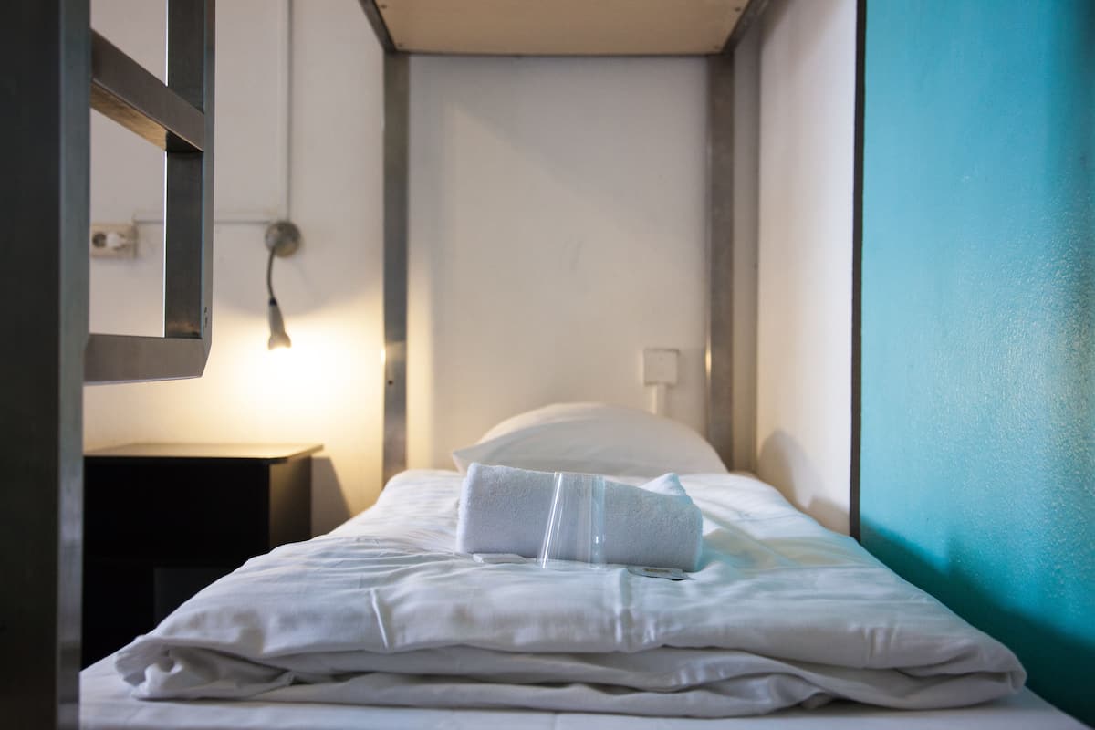 A neatly made bed with a pillow and a folded towel, a bedside table with a lamp, and a window with a view of the outside.