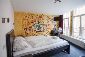 A hotel room with a large bed, a wall mural, a window with curtains, a bedside table with a lamp, and a flat-screen TV. - St Christopher's at The Winston - Hostel (Amsterdam)